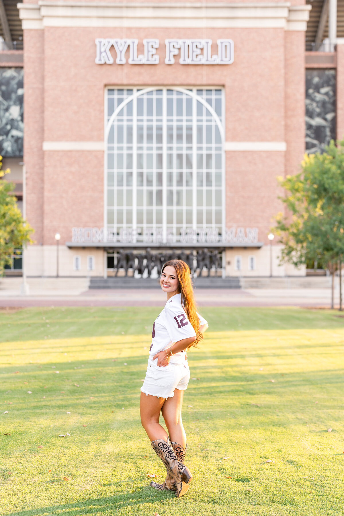 Texas A&M senior girl standing with hands in back pocket and smiling over shoulder in front of Kyle Field at Aggie Park while wearing white jersey, shorts and boots