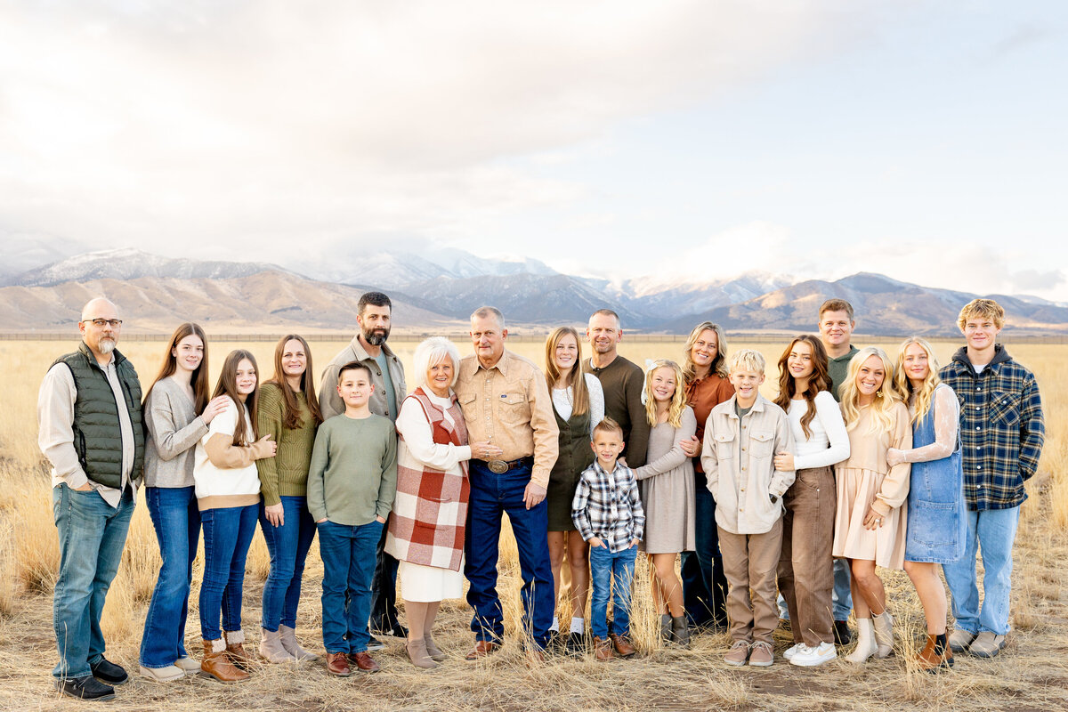 A large extended family poses together in a vast open field in Eagle Mountain, Utah, with snow-capped mountains in the distance. They are dressed in coordinated neutral and earth-toned outfits, capturing a timeless family moment.
