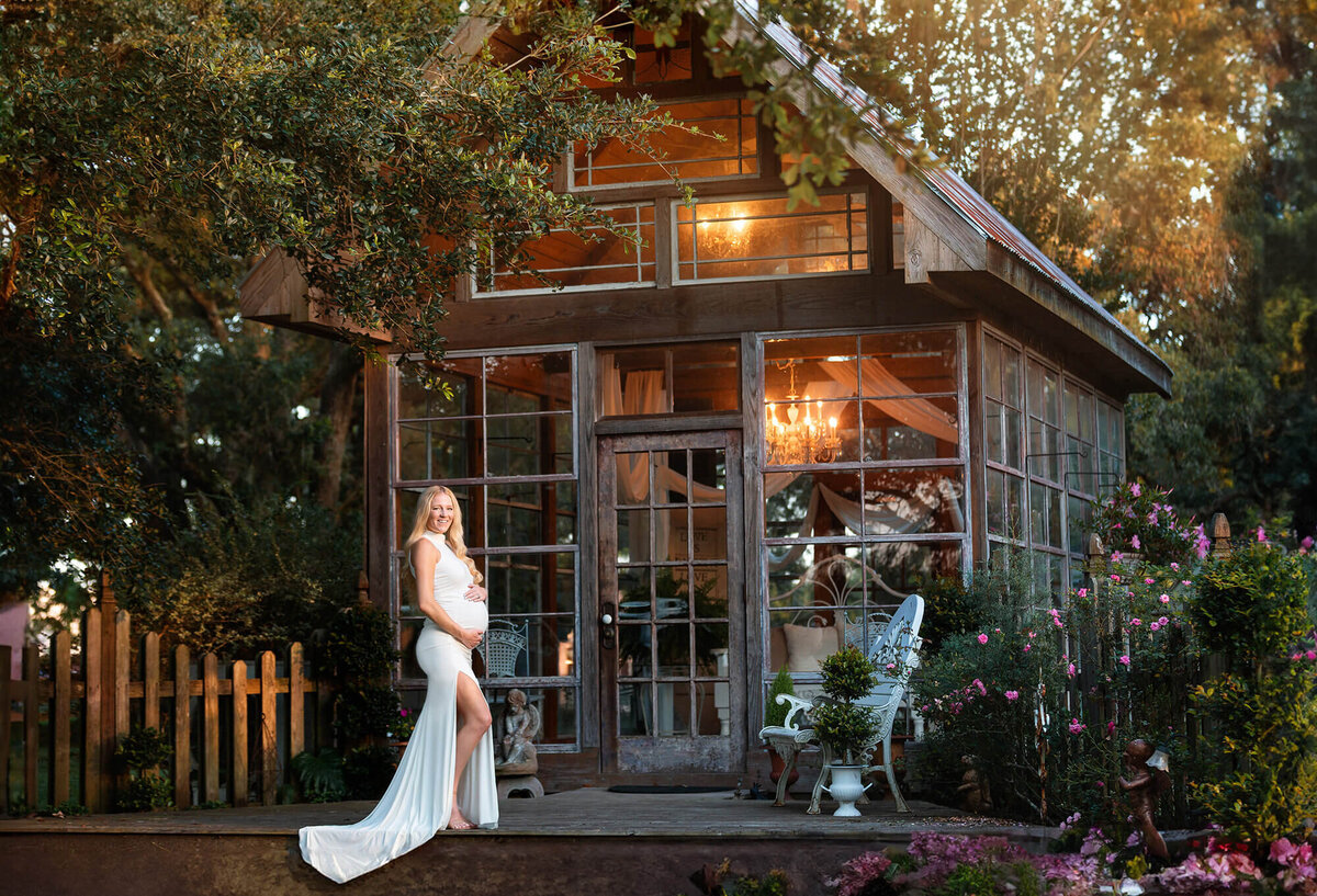Gorgeous blonde mom to be wearing a pure white dress in front of a greenhouse in the beautiful location in Angleton called Out Under the Trees.