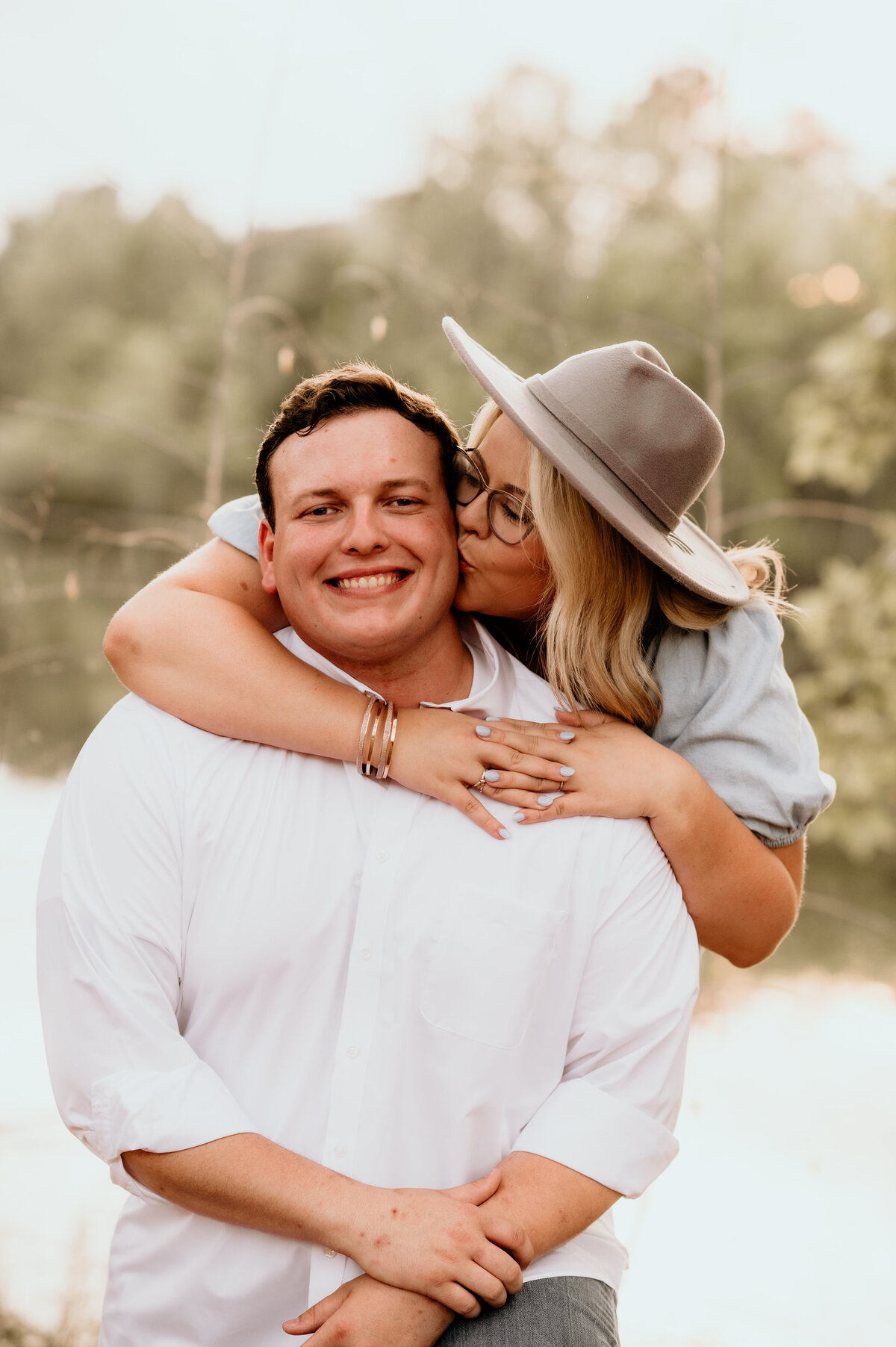 woman standing behind a man who is sitting in front of a lake, the woman wraps her arms around the man and kisses his cheek for their outdoor engagement session