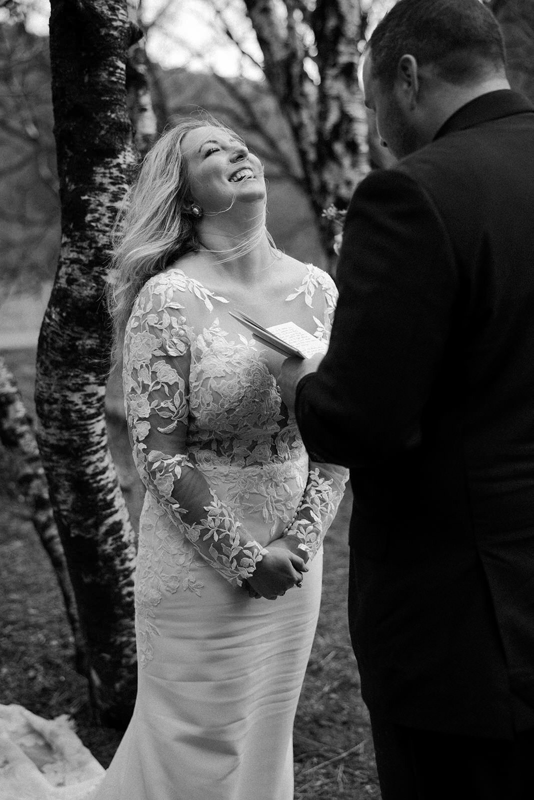 A heartfelt black and white photo of a bride laughing during a vow reading in a forested setting