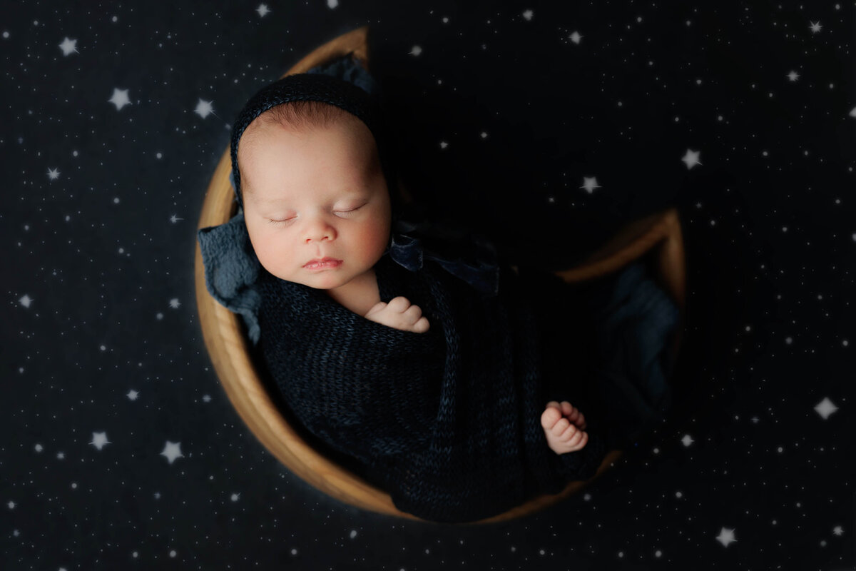 newborn baby in a blue swaddle and bonnet in a wooden moon bowl on a blue backdrop with stars at his newborn photography session with a photographer at a chantilly photo studio