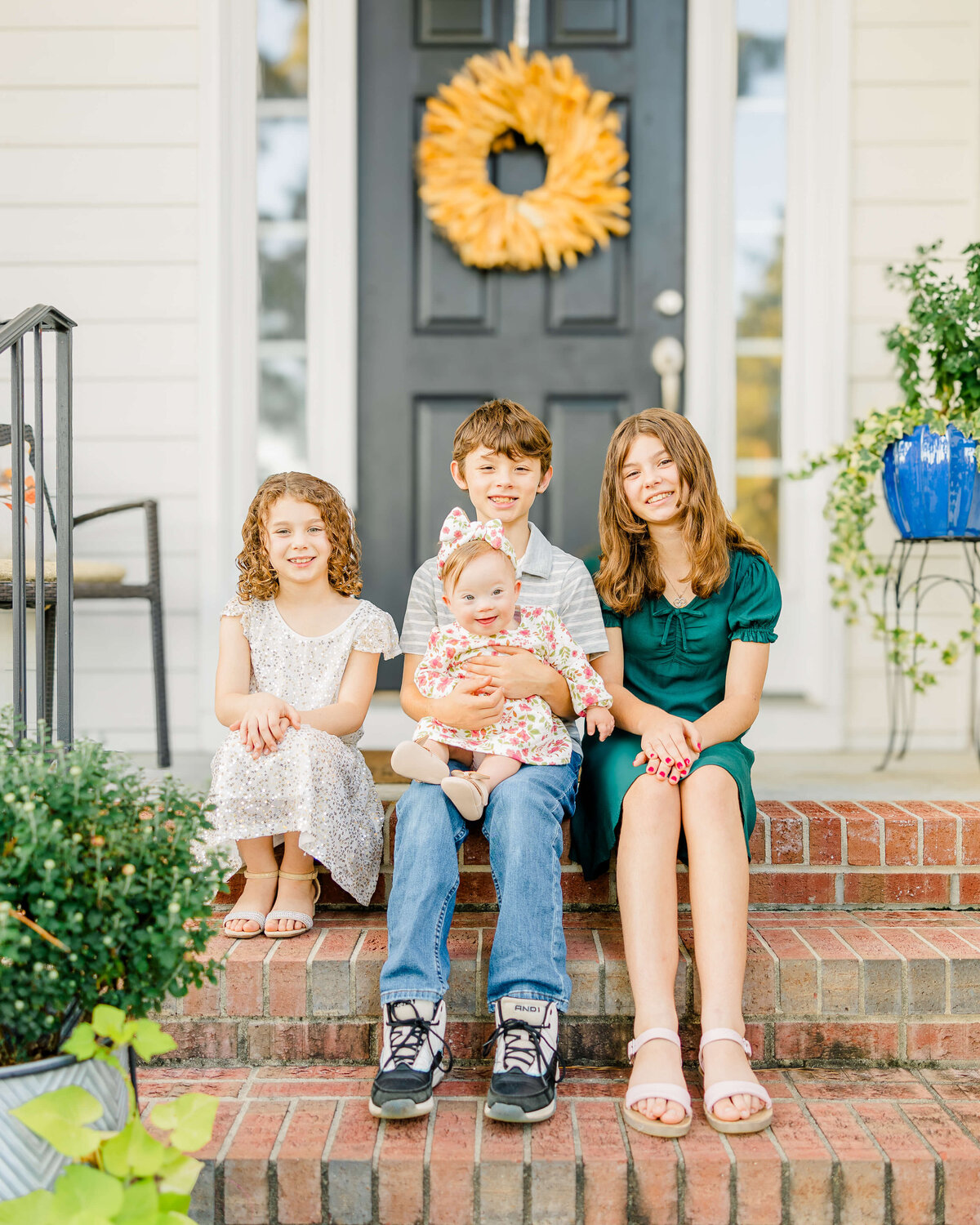 Four children sit on brick steps in front of a black door adorned with a yellow wreath. The older three kids smile while the middle child holds a baby in a floral outfit. Captured by a Fayetteville, NC family photographer, potted plants flank the steps, and the home exterior is off-white.