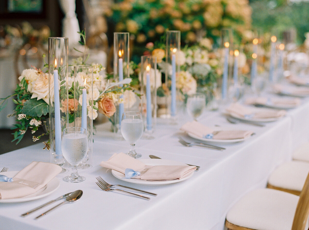 Elegant long tablescape with blue taper candles in glass holders and lush floral centerpieces in blush, ivory, and peach.