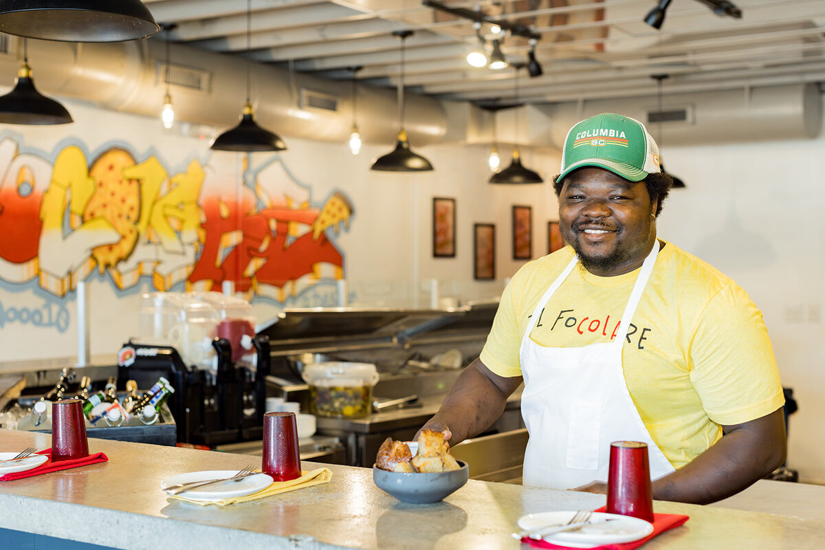 Smiling restaurant owner in Columbia, SC, wearing a yellow t-shirt, white apron, and Columbia SC hat, presenting a bowl of fresh bread behind the counter. The modern eatery features industrial lighting and a graffiti-style mural. Small business branding photography in Columbia, SC, capturing a local entrepreneur in the food industry.