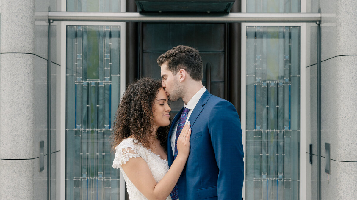 A bride and groom stand facing each other in front of a temple of the Church of Jesus Christ of Latter-Day Saints and the groom kisses the bride on the forehead while the bride rests her hand on the grooms chest