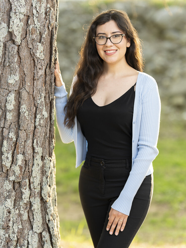 Woman wearing black jeans and a black top with a light blue sweater stands next to a tree