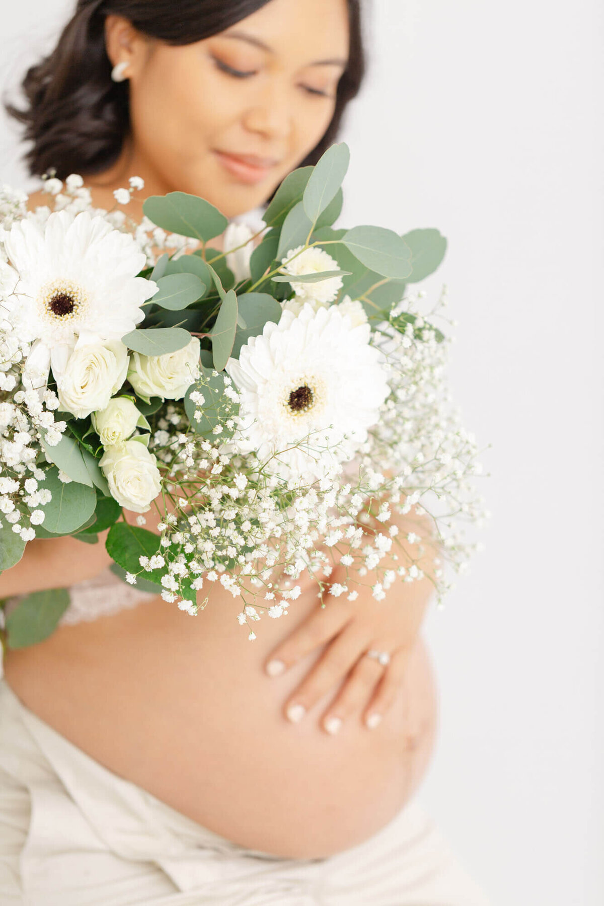 Pregnant Mama holding a bouquet of white flowers in front of her. The flowers are in focus and her face and baby bump are a little softer in the background. She has one hand on her belly.