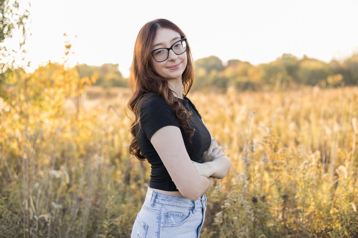 High school senior female, with long black hair, wearing glasses, blue jeans, short sleeve black shirt, standing in grass with trees in the background, turned to the side, smiling at the camera