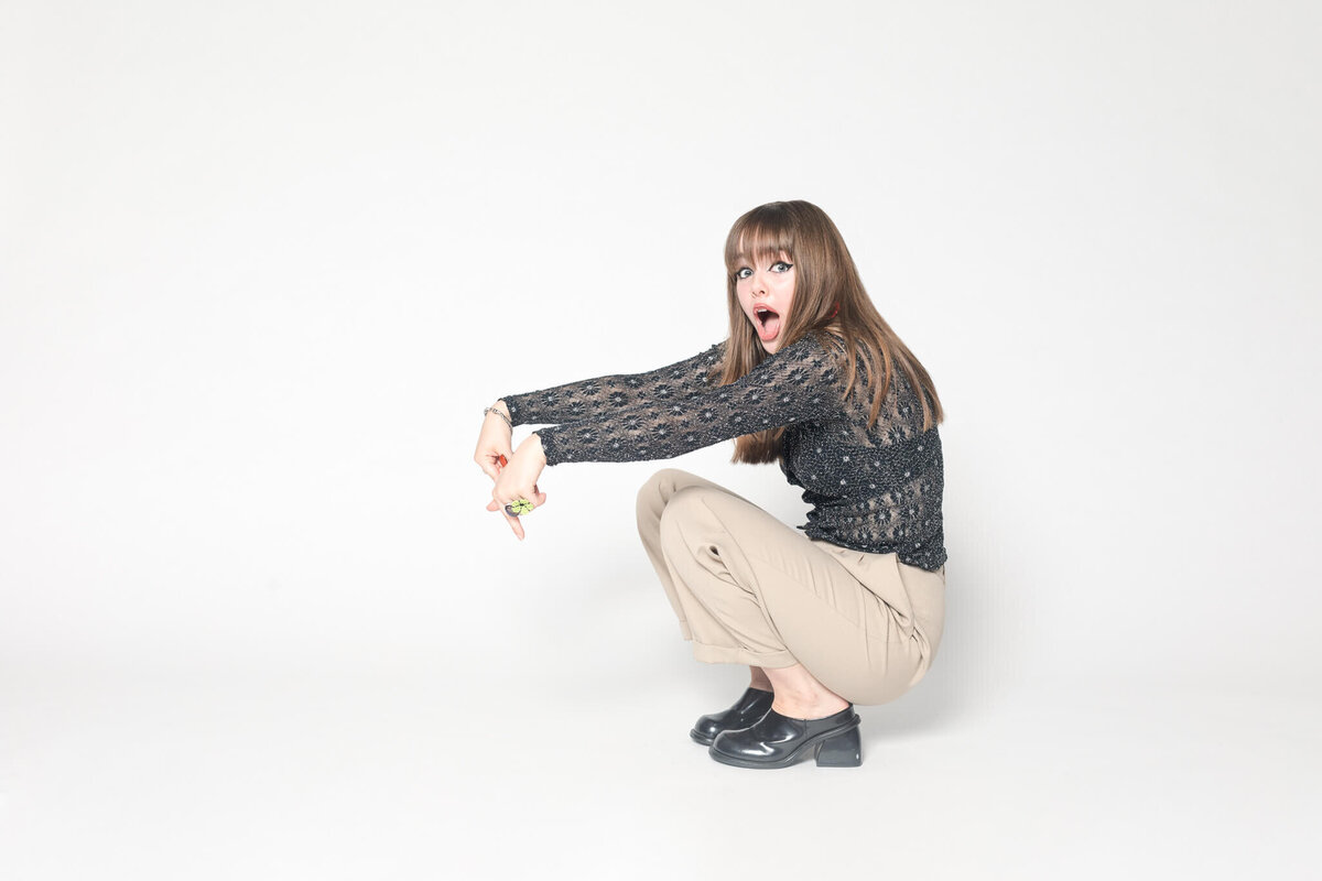A woman with long hair, wearing a black patterned top and beige pants, squats with a surprised expression against a plain white background. She points downward with both hands.