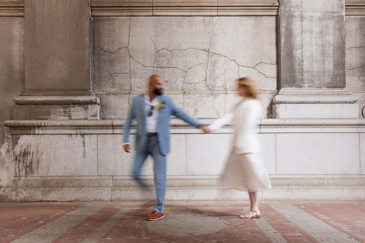 Blurred motion photo of a couple holding hands while walking in front of the stone walls of the NYC Marriage Bureau, styled in light wedding attire—an artistic image by an NYC elopement photographer that blends city architecture with candid movement.