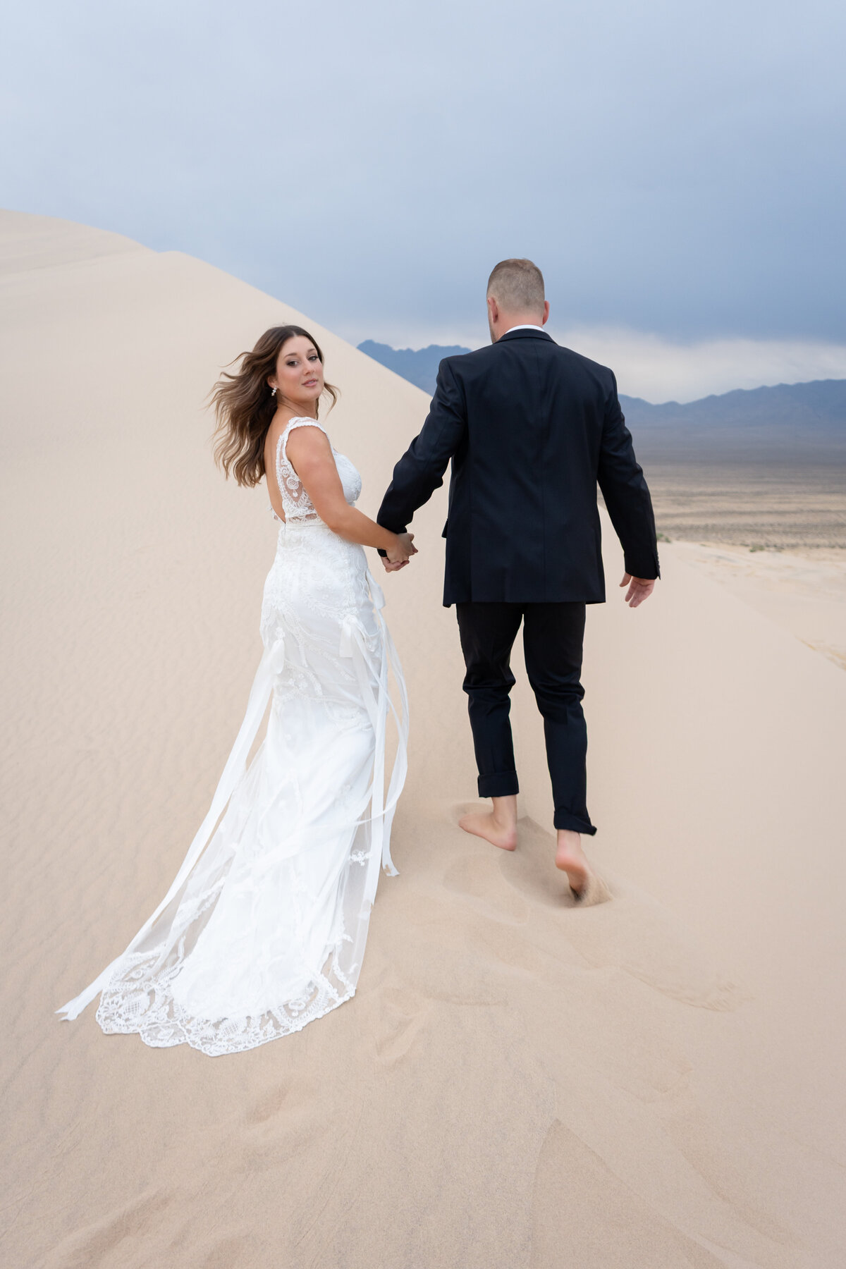 Bride and groom at Kelso Dunes at sunrise walking together holding hands while the bride is wearing a Claire Pettibone lace gown with ribbons