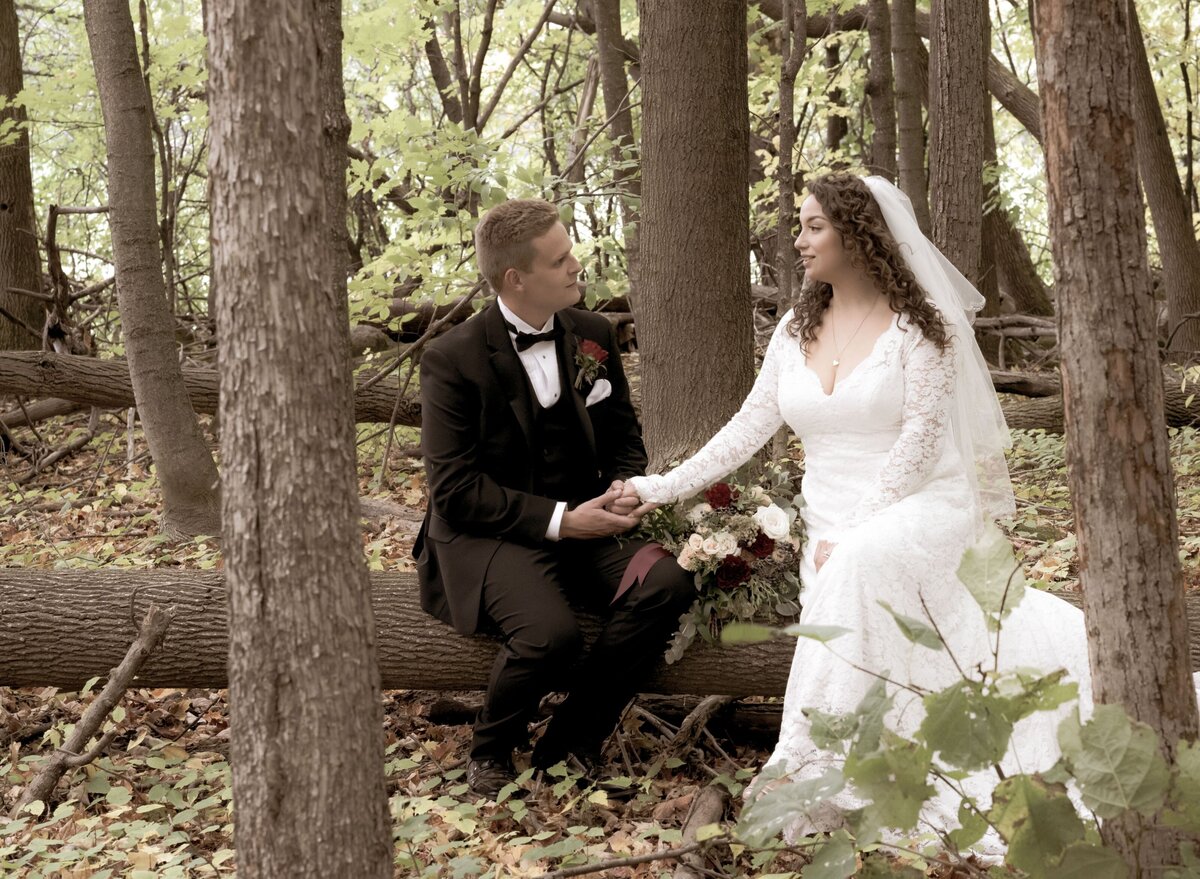 The bride and groom sit together on a log in a serene forest setting. Surrounded by lush greenery, they enjoy a peaceful and intimate moment, highlighting their connection and the natural beauty of their environment.