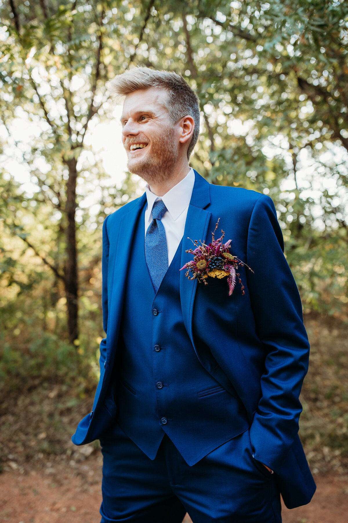 Groom in a bright blue suit smiling confidently in a forest setting, with a colorful boutonniere