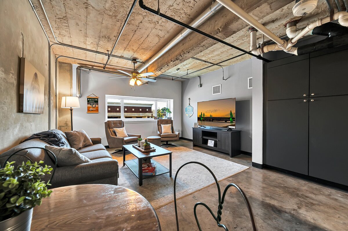 Open concept living room area with comfortable seating and smart TV in this one-bedroom, one-bathroom industrial vacation rental condo with free Wifi, onsite laundry, and space for four in the historic Behrens building of downtown Waco, TX.