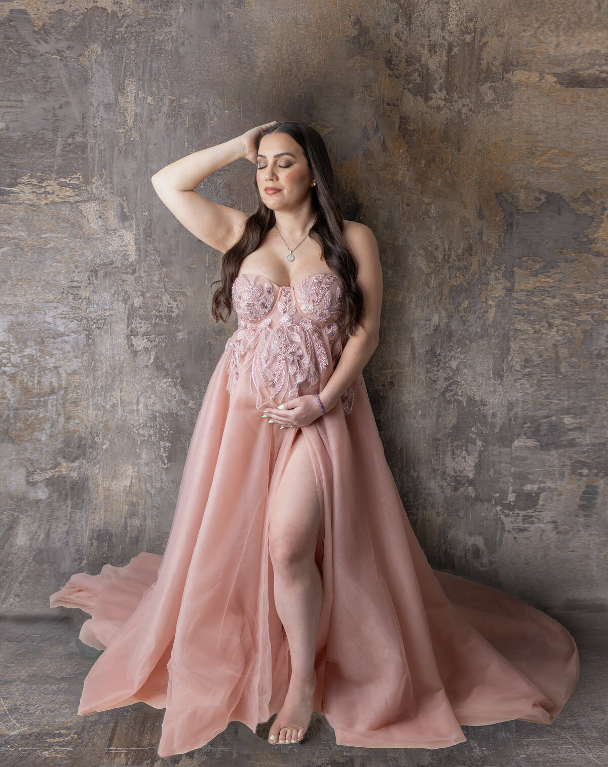 Pregnant woman in a flowing blush gown standing gently holding her belly and smiling softly during a maternity shoot in Waldwick, NJ.