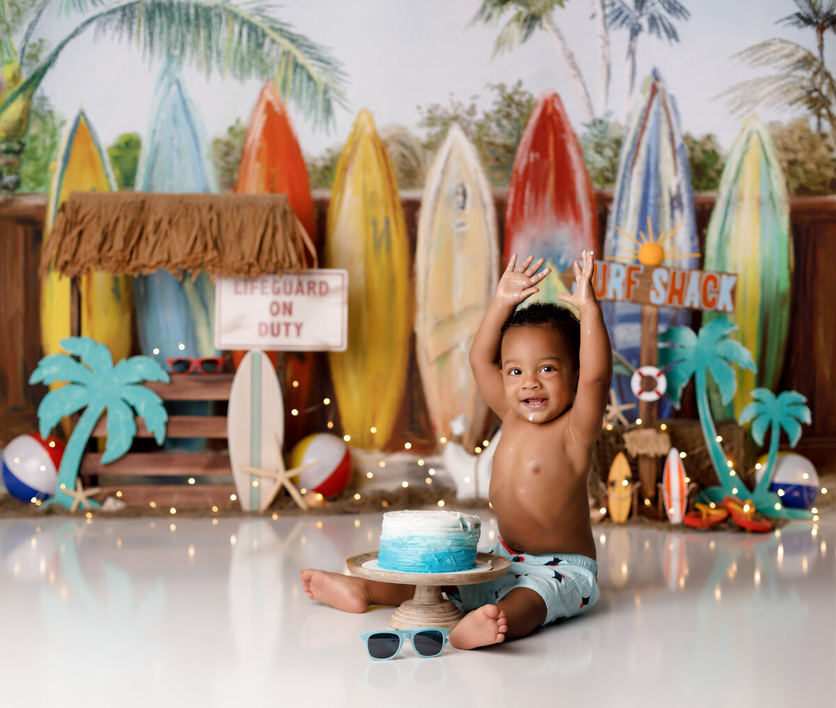 Surf and beach theme cake smash in Palm Beach and Boca Raton newborn and cake smash portrait studio. Baby boy is wearing light blue swim trunks sitting in front of an ombre Blue cake. In the background are various surfboards with a rustic tiki hut and surf shack sign. There are aqua palm trees and beach balls on the ground.