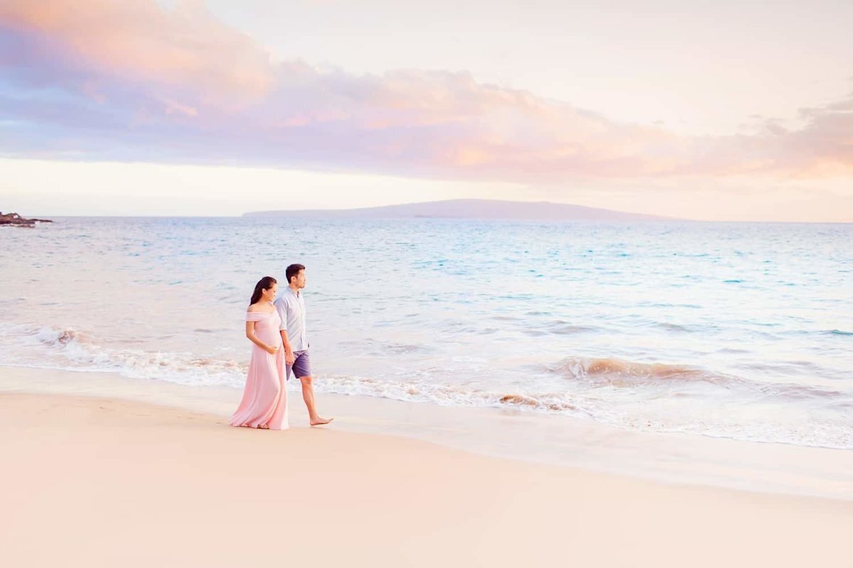 Beautiful sunrise light surrounds a couple walking hand-in-hand on the beach in Wailea as Love + Water photographs their Maui babymoon