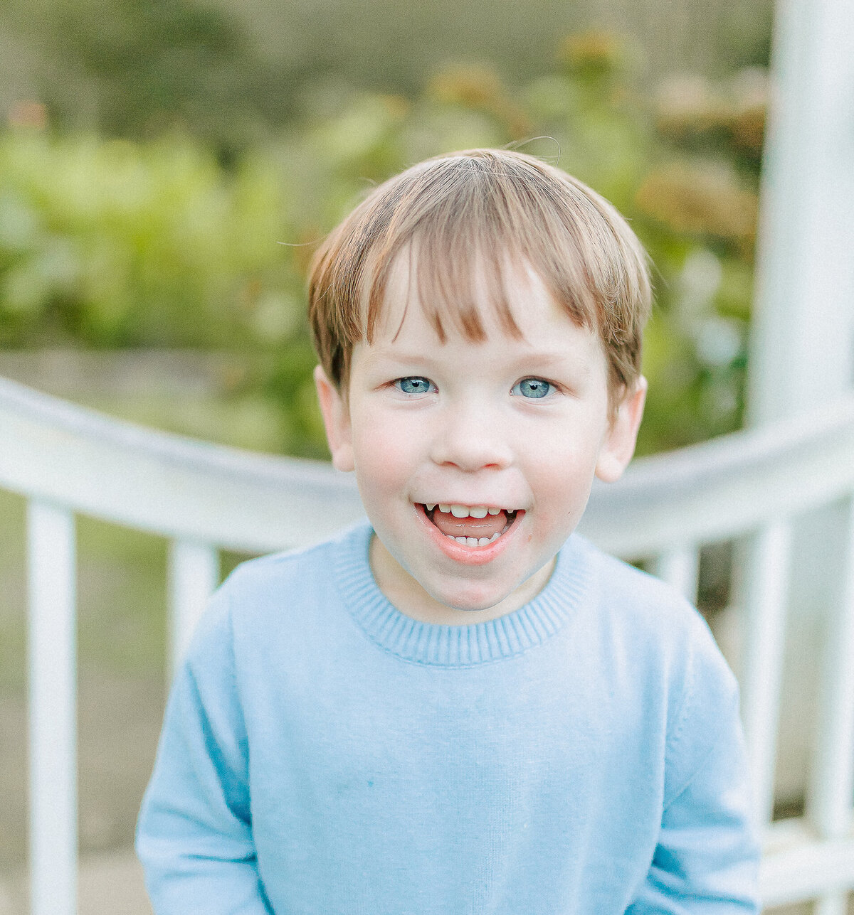 A young boy with bright blue eyes and a joyful smile, wearing a light blue sweater against a soft, outdoor backdrop.