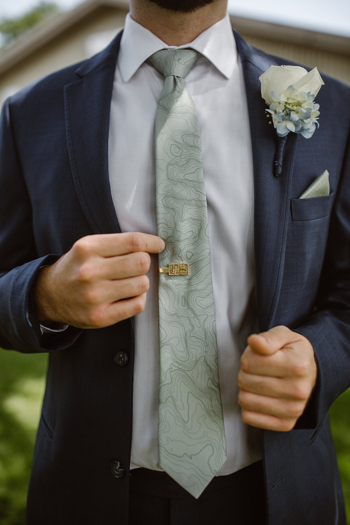 Close-up of a groom adjusting his patterned tie with a custom gold tie clip, wearing a navy suit, white shirt, and boutonniere with a white rose and blue hydrangea.