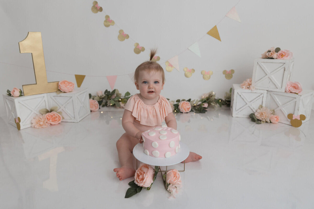 Baby girl sitting in front of a pink polka dot cake, surrounded by floral decorations and a gold number one sign for her first birthday photoshoot.