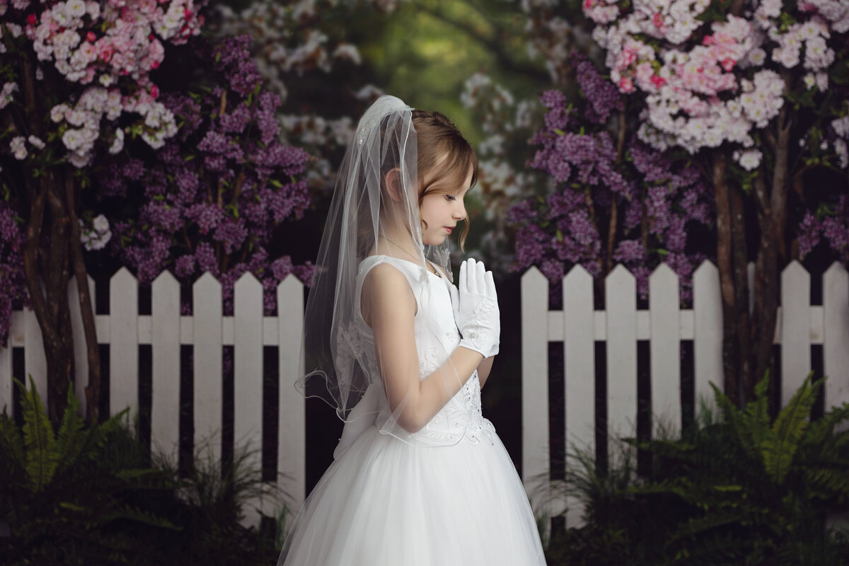 A young girl prays with white gloves and dress in a studio of a New Jersey Communion Portrait Photographer in front of a white picket fence and blooming trees