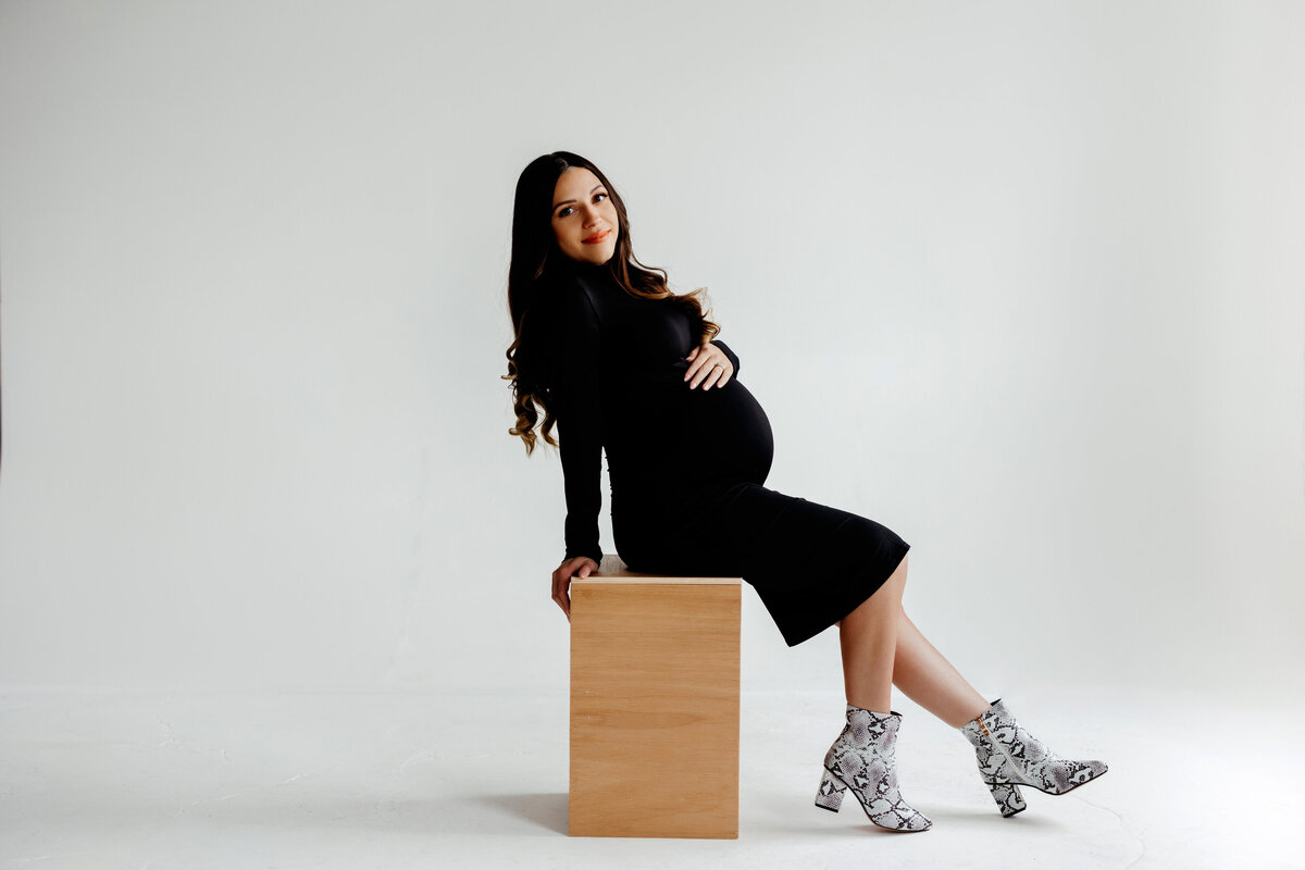 pregnant mom wears a tight black dress while sitting on brown posing block