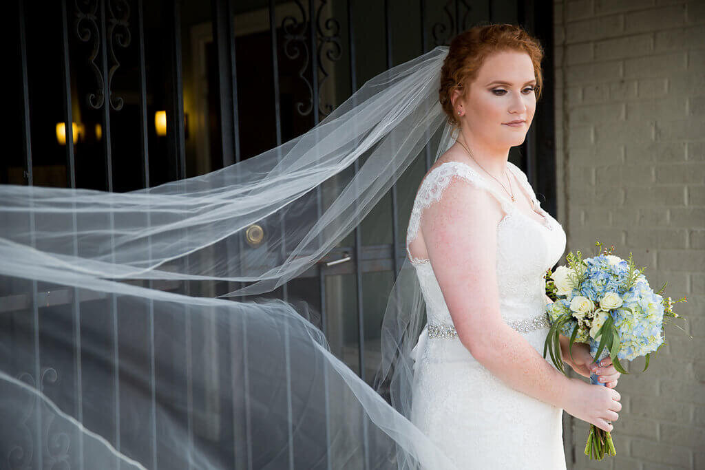 A bride in a white lace wedding dress and long veil stands holding a bouquet of blue and white flowers, looking to the side with a soft expression. The veil flows gently behind her.