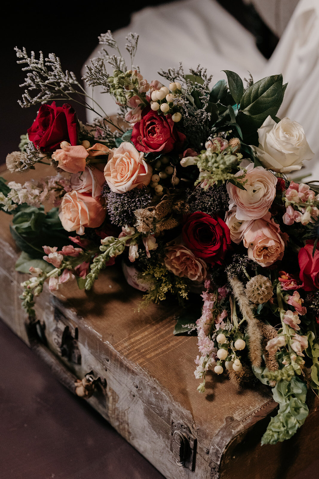 A close-up of the bride’s lush bouquet, featuring red, peach, and white roses, greenery, and berry accents, resting on a vintage suitcase.