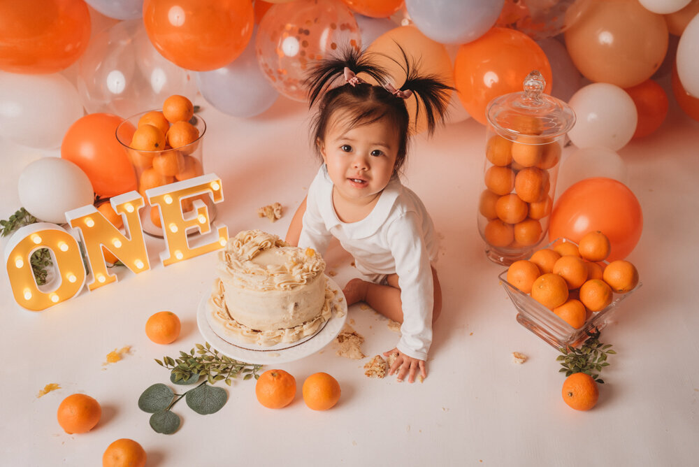 Photo shoot with Atlanta, Georgia cake smash photographer doing a clementine themed portrait session for baby girl's one year birthday with baby sitting on white backdrop next to birthday cake surrounded by clementine's, greenery, light up O N E marquee letters and orange balloon garland behind her. Baby is smiling looking up at camera