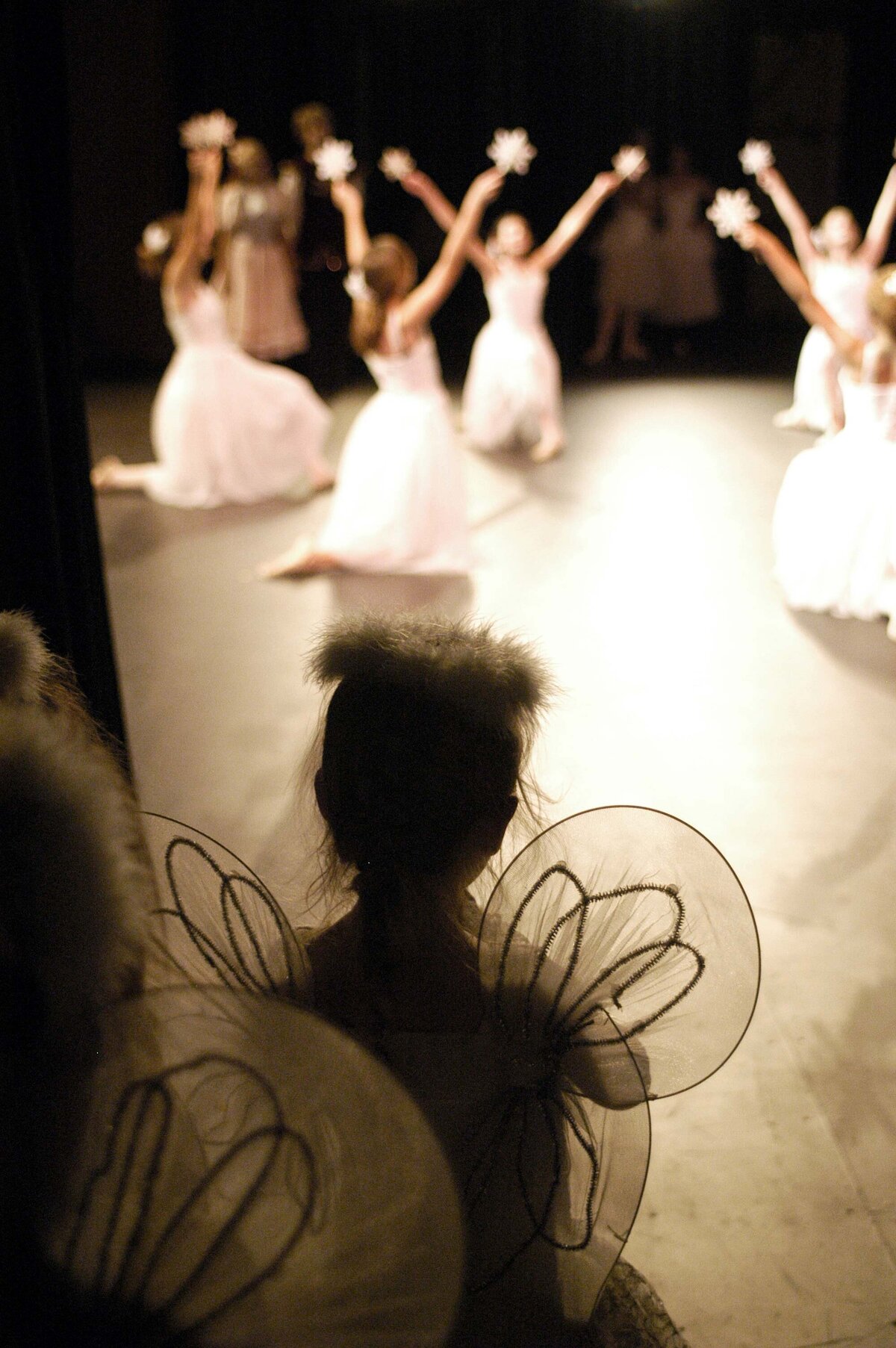 A little dancer watches older modern dance performers from the wings. She wears angel wings and is silhouetted by the stage light to imply a story