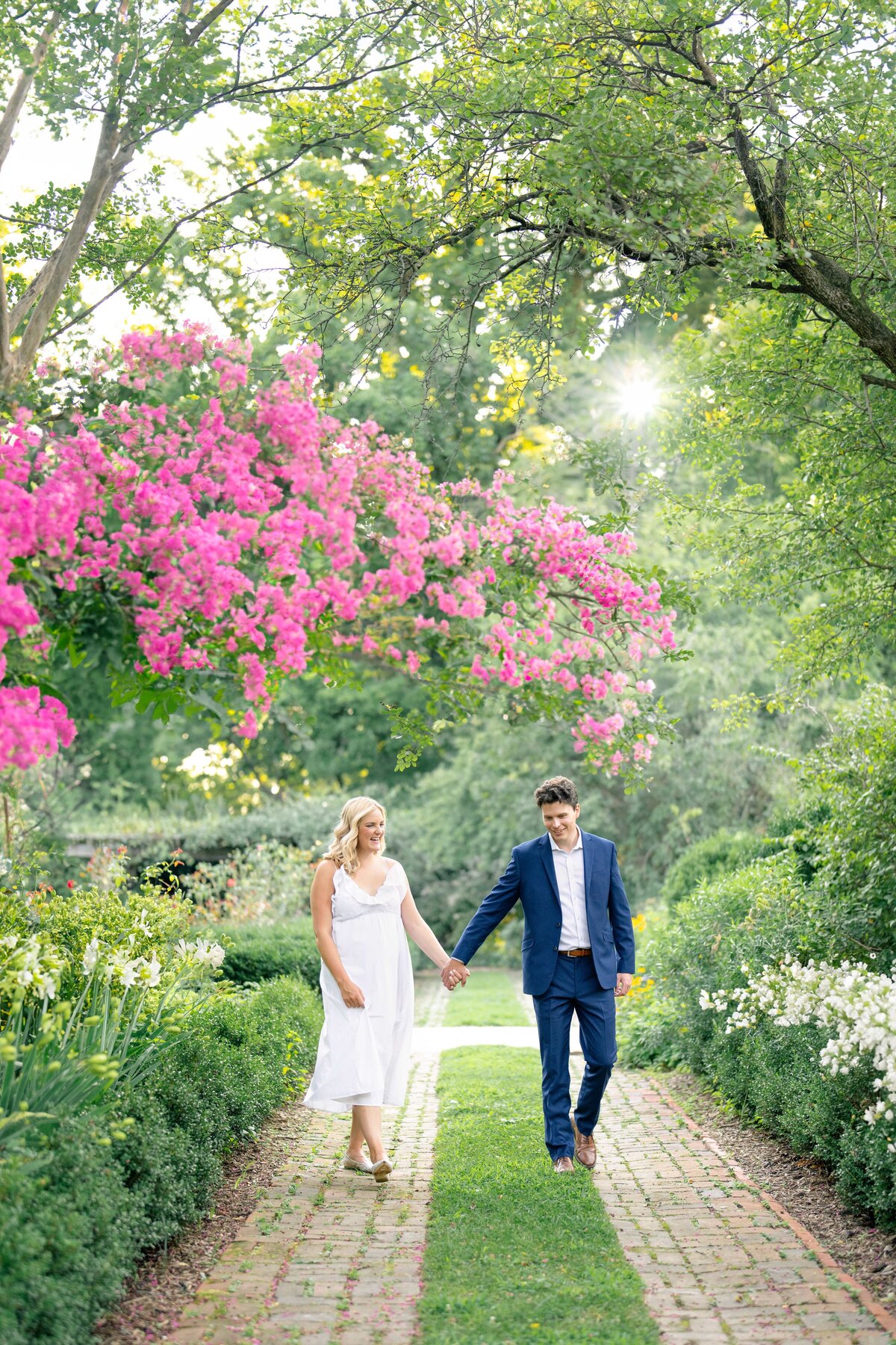 A couple holding hands walks along a brick path in a garden. The man is in a blue suit, and the woman is in a white dress. They are surrounded by greenery and vibrant pink flowers overhead, with sunlight filtering through the trees.