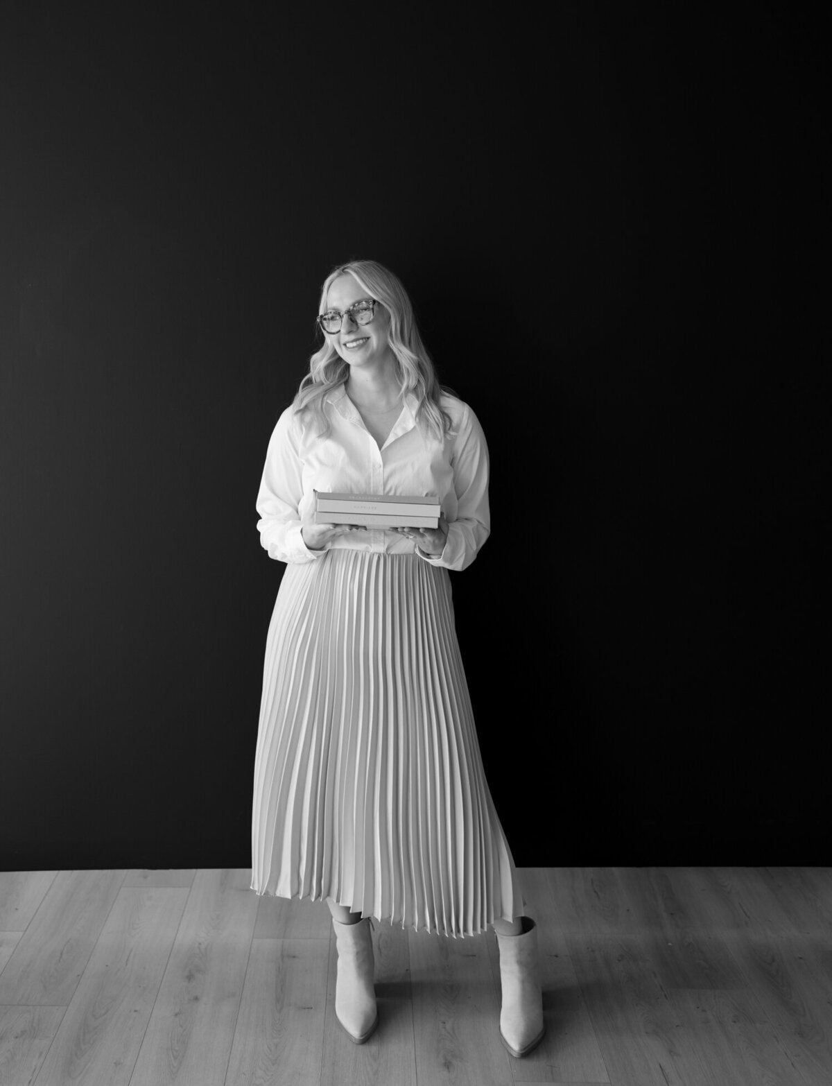 blonde woman in champagne pleated skirt holds linen journals with a black studio backdrop