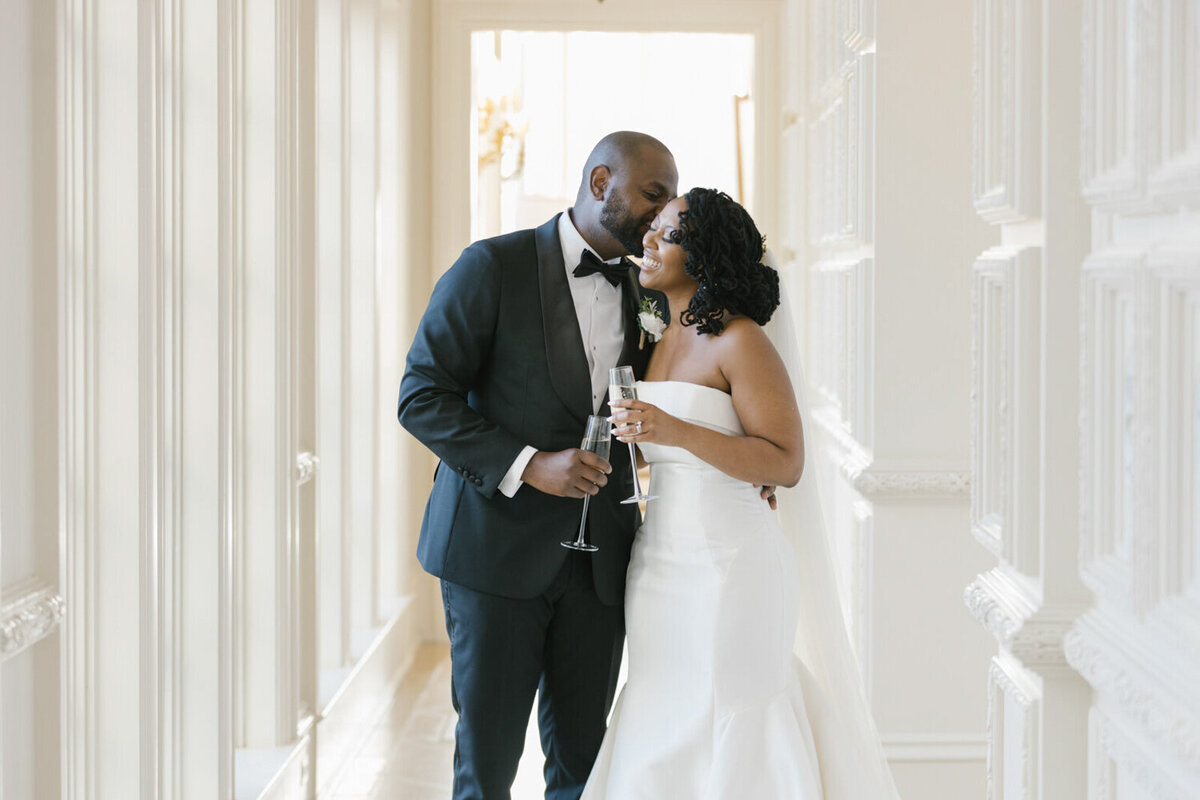 Bride and groom giggling while holding champagne in the white European-style hall at The Olana wedding venue.
