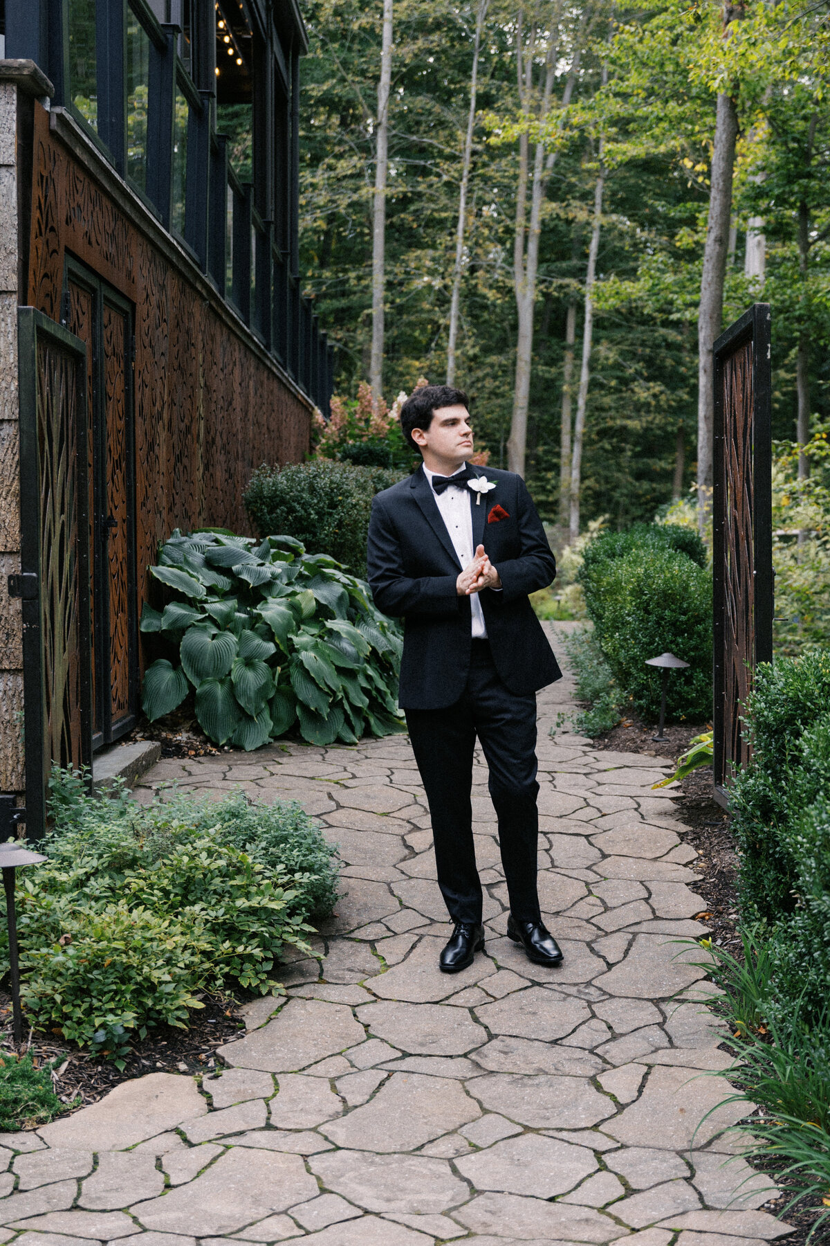 Groom standing in garden walkway at Sapphire Creek Winery, black tux and red boutonnière

