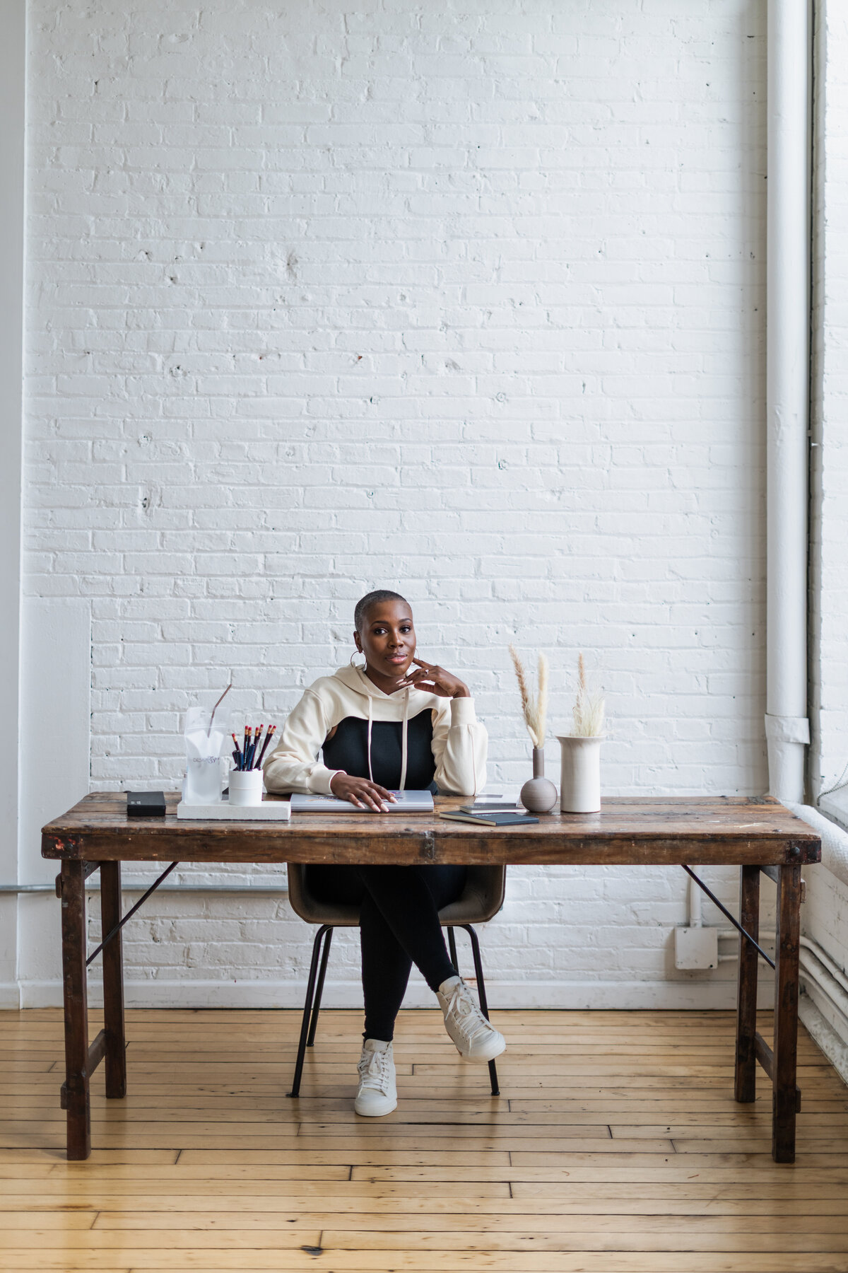 A branding image of a creative entrepreneur at her wooden desk, framed by white brick walls and warm natural light. The scene reflects her focus and the intentional workspace behind her acrylic signage business.