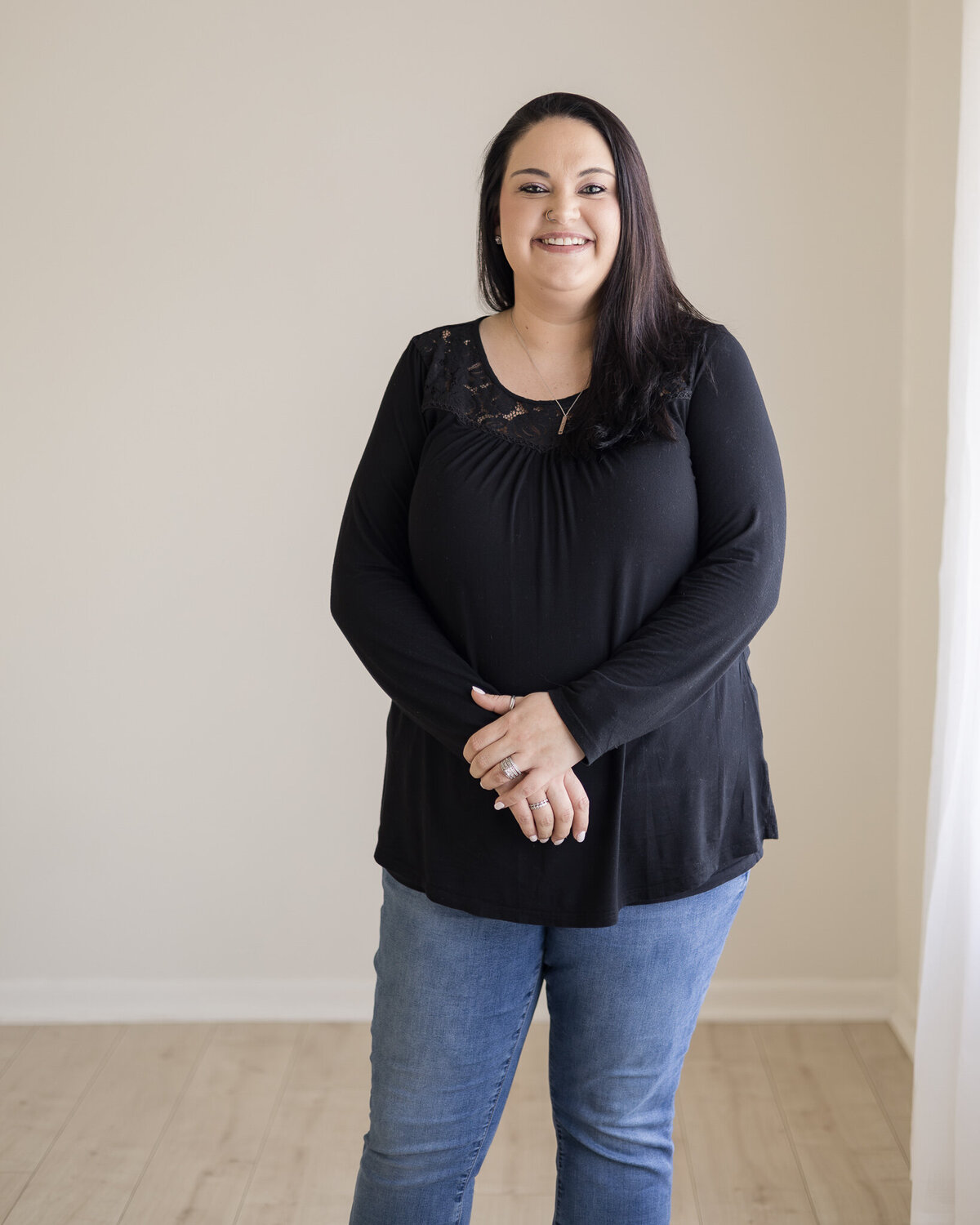 Woman standing, has black hair, wearing a black shirt, blue jeans, and black shoes, smiling at the camera