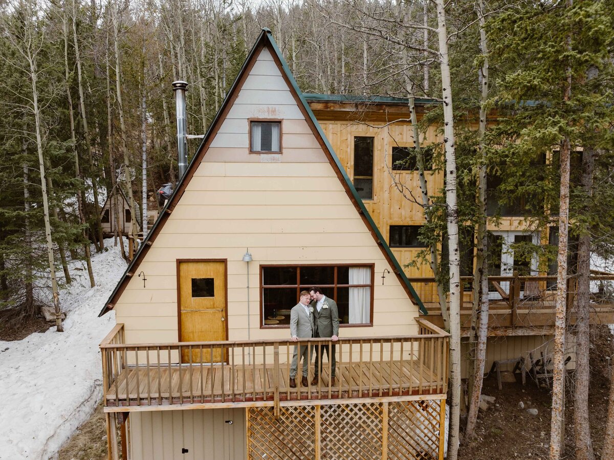 Two grooms share an intimate moment on the balcony of an A-frame cabin surrounded by aspen and pine trees during their Copper Mountain, Colorado elopement.