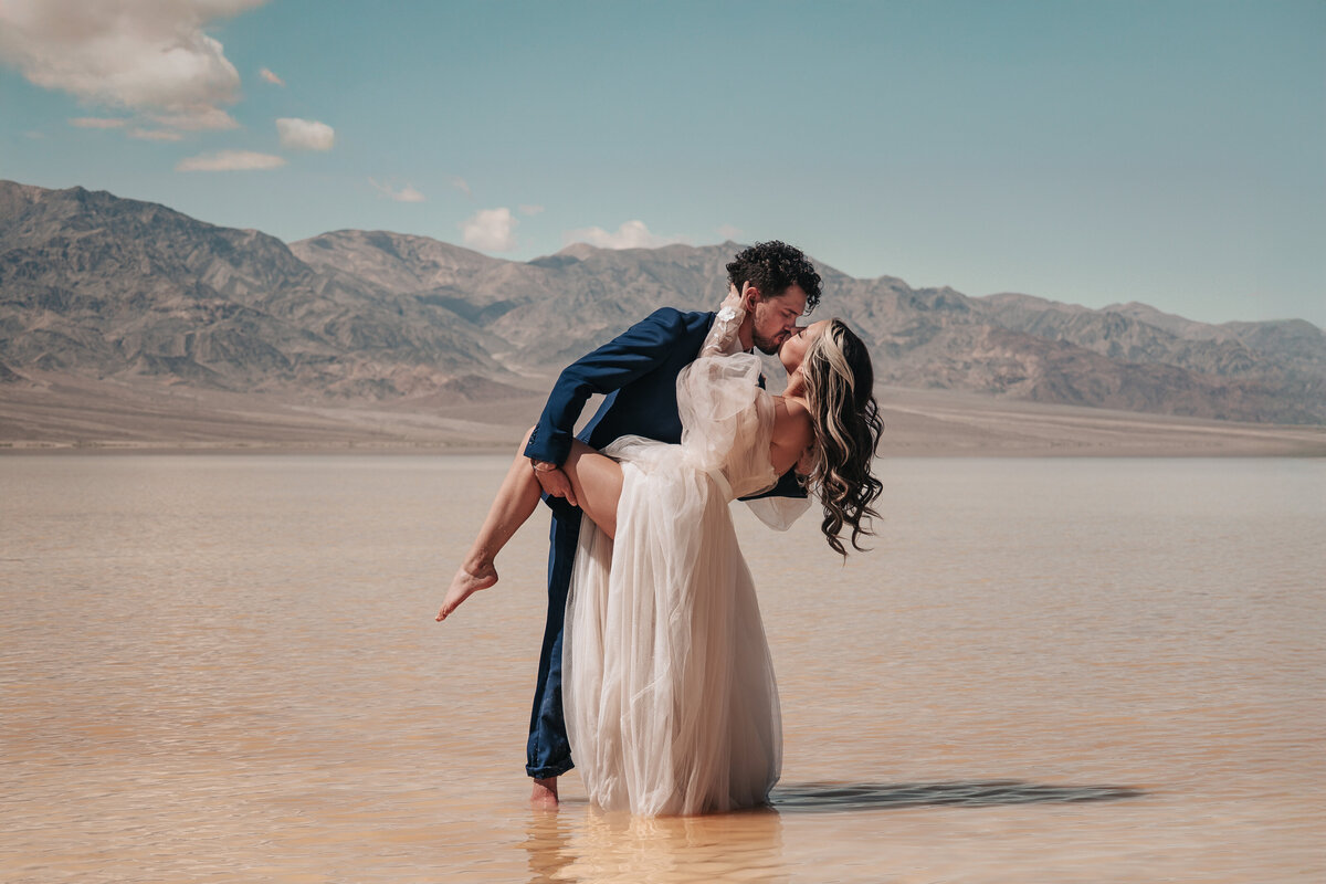 Fashion-forward wedding couple engages in a passionate dip kiss at Death Valley's Bad Water Basin featuring the dramatic desert mountain landscape, captured by a destination photographer specializing in bold and artistic bridal sessions.