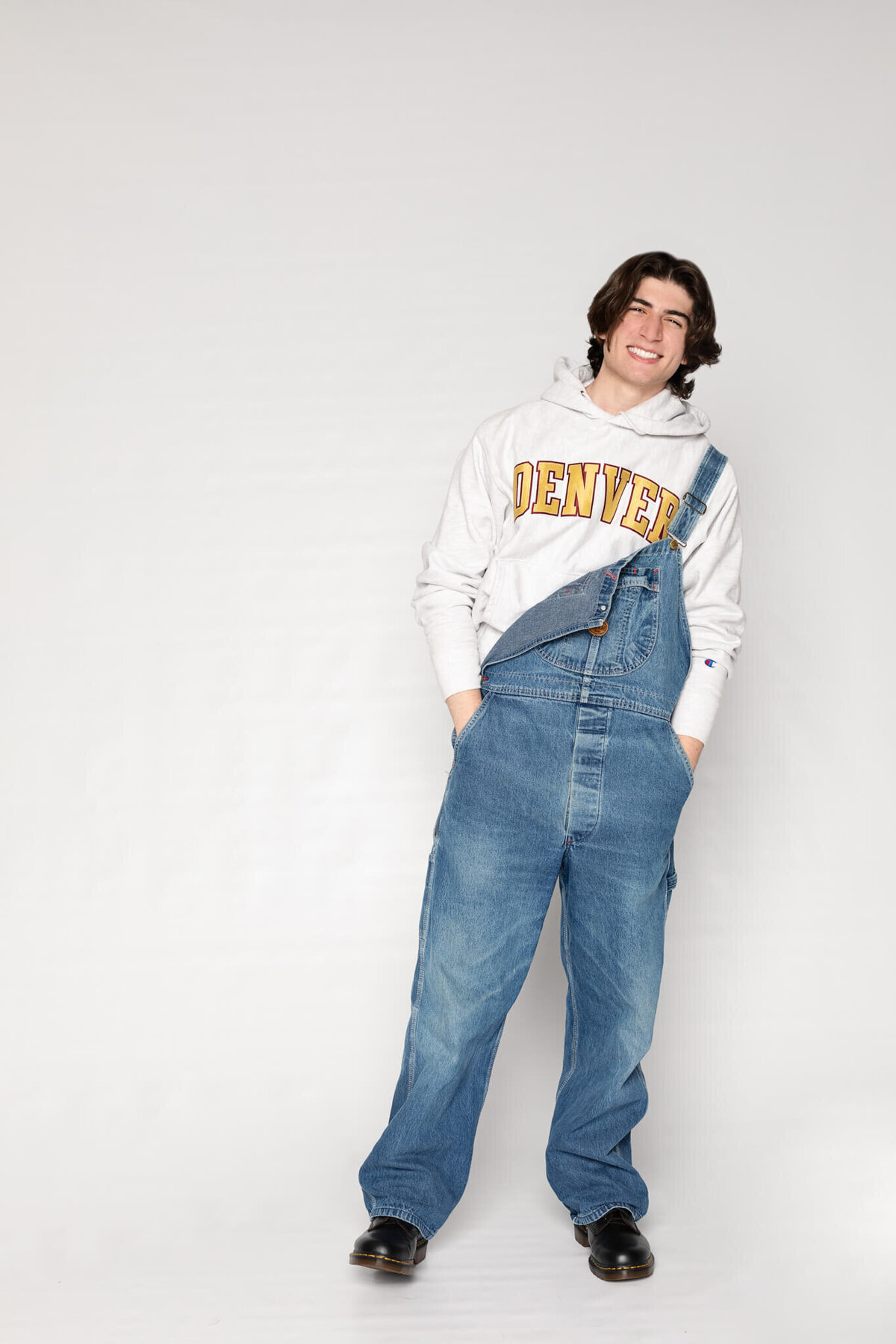 A person with long hair smiles while standing and leaning slightly. They are wearing a Denver sweatshirt underneath denim overalls and black shoes. The background is plain and light gray.