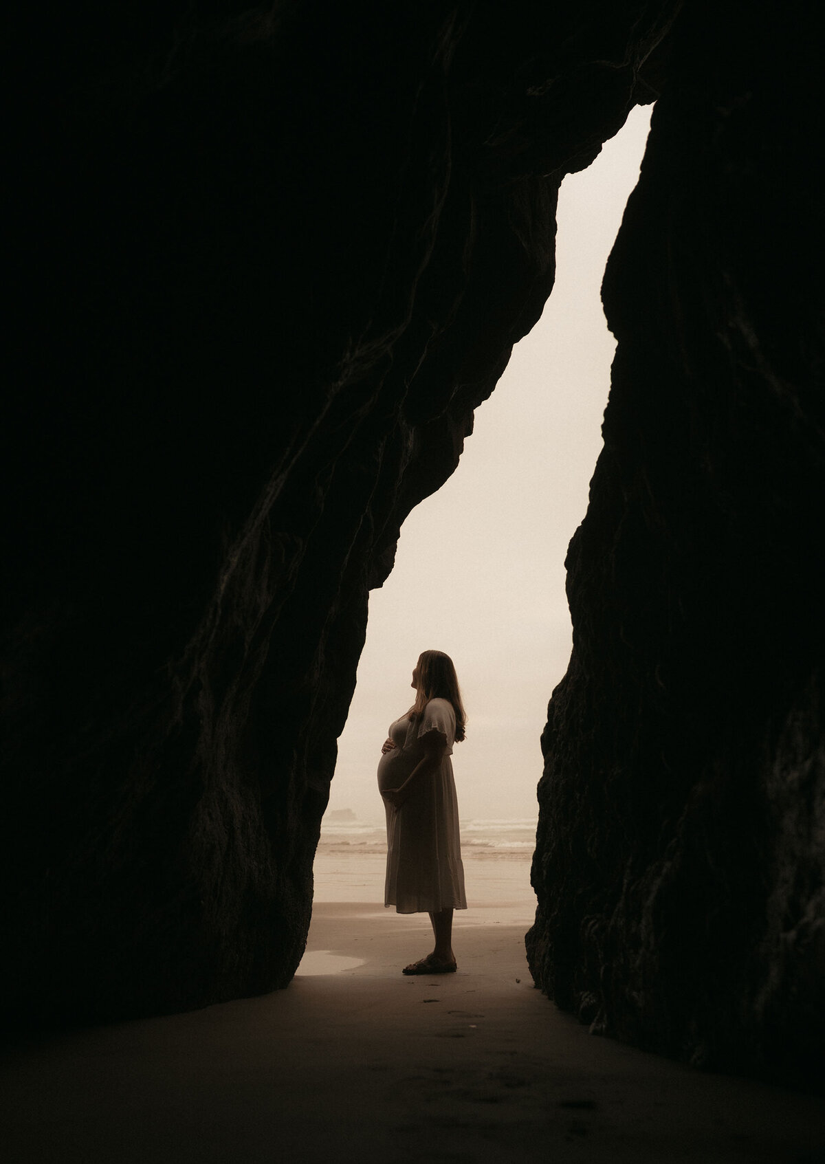 pregnant woman stands against beachy backdrop with her hand on her belly at the Oregon Coast