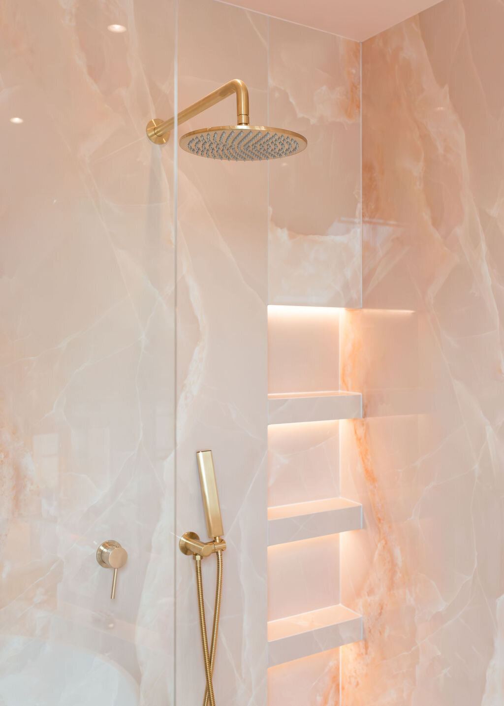 A luxurious glass-enclosed walk-in shower, featuring pink marble walls, gold hardware, built-in shelving, and a bench.