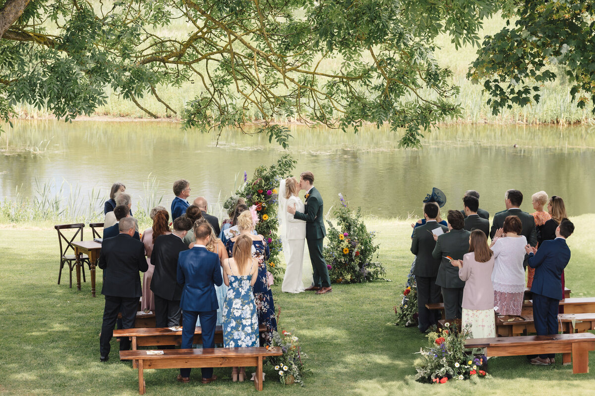 A romantic moment during the wedding ceremony at Willow Marsh Farm, as the bride and groom share their first kiss. They stand beneath a beautiful tree, with vibrant floral arrangements on either side of them, while guests watch on from wooden benches. The serene pond in the background adds to the peaceful, intimate atmosphere. Guests are clapping and celebrating as the couple enjoys this unforgettable moment.