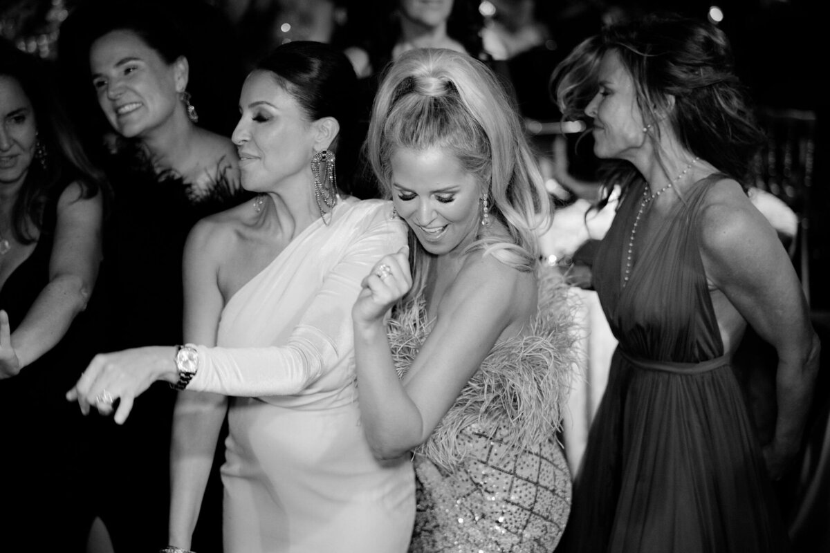 The lively event or party captured in the black and white photo took place at Virginia, specifically at a tented wedding photographed by Elisabetta Marzetti, with Clayton Films as videographers. In the center of the image, a group of women in elegant evening attire can be seen dancing and smiling. One particularly striking woman stands out in her sparkling dress adorned with feathers, adding to the overall vibrancy and joy captured in the scene.   Is there anything else you would like to add or modify?