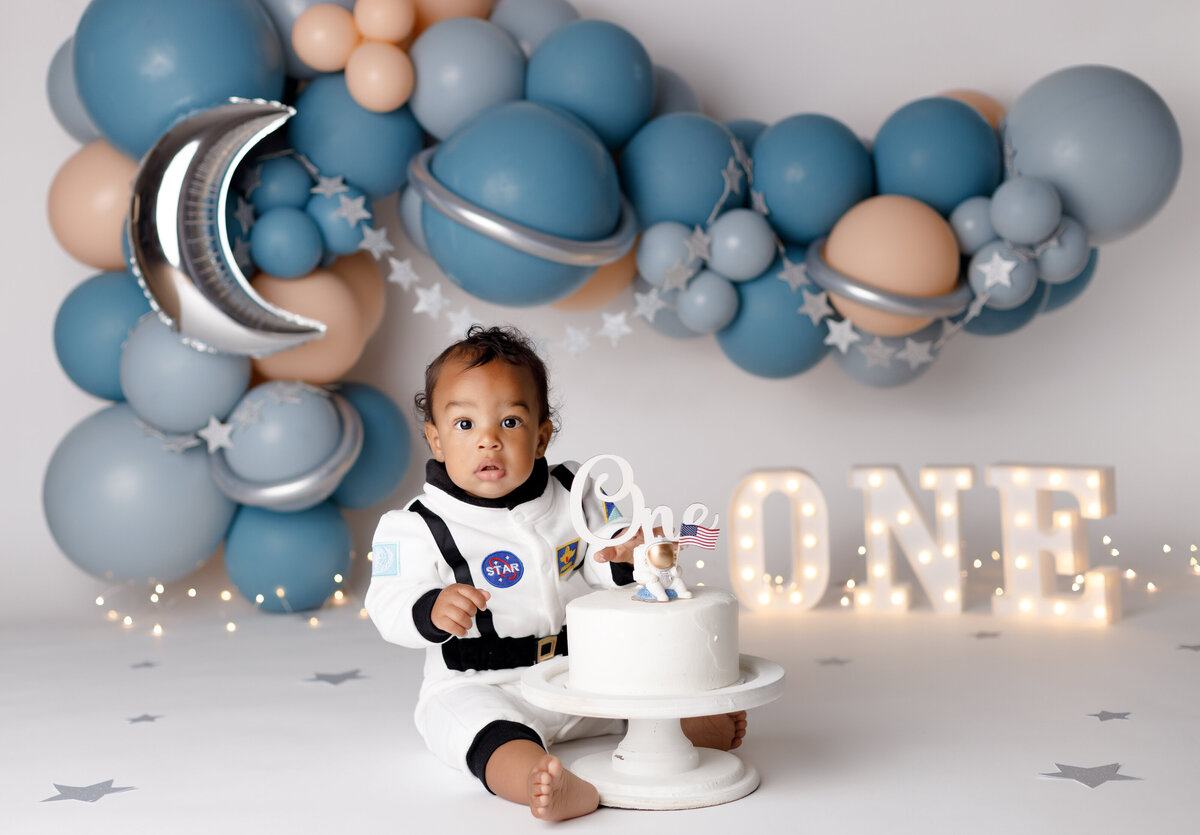 Space themed cake smash in West Palm Beach and Boca Raton newborn and cake smash photography studio.  Baby is dressed in an astronaut outfit sitting behind a white cake.  In the background is a space theme balloon garland with star garland and a light up one.