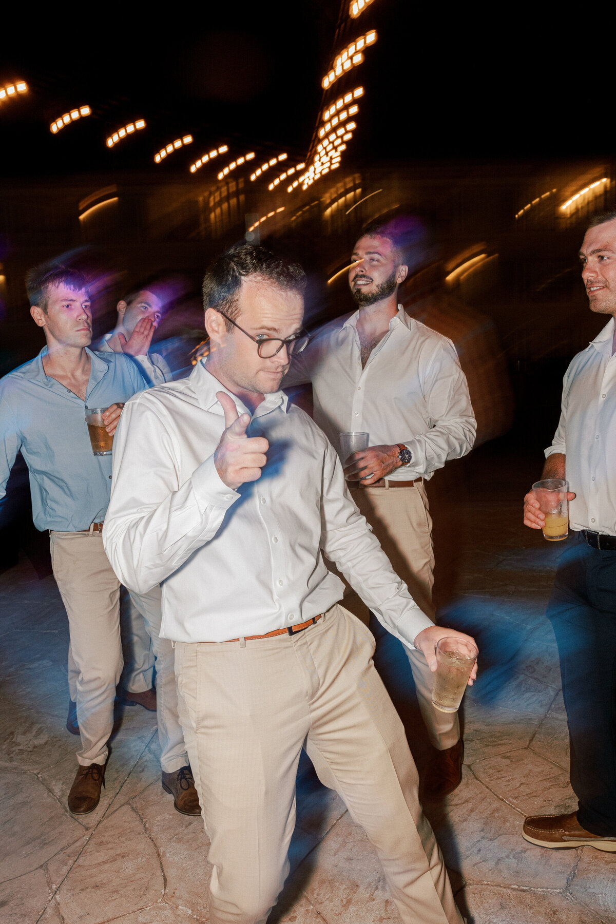Groomsman dances under string lights at a Cancun wedding reception, drink in hand, captured by Philly photographer.