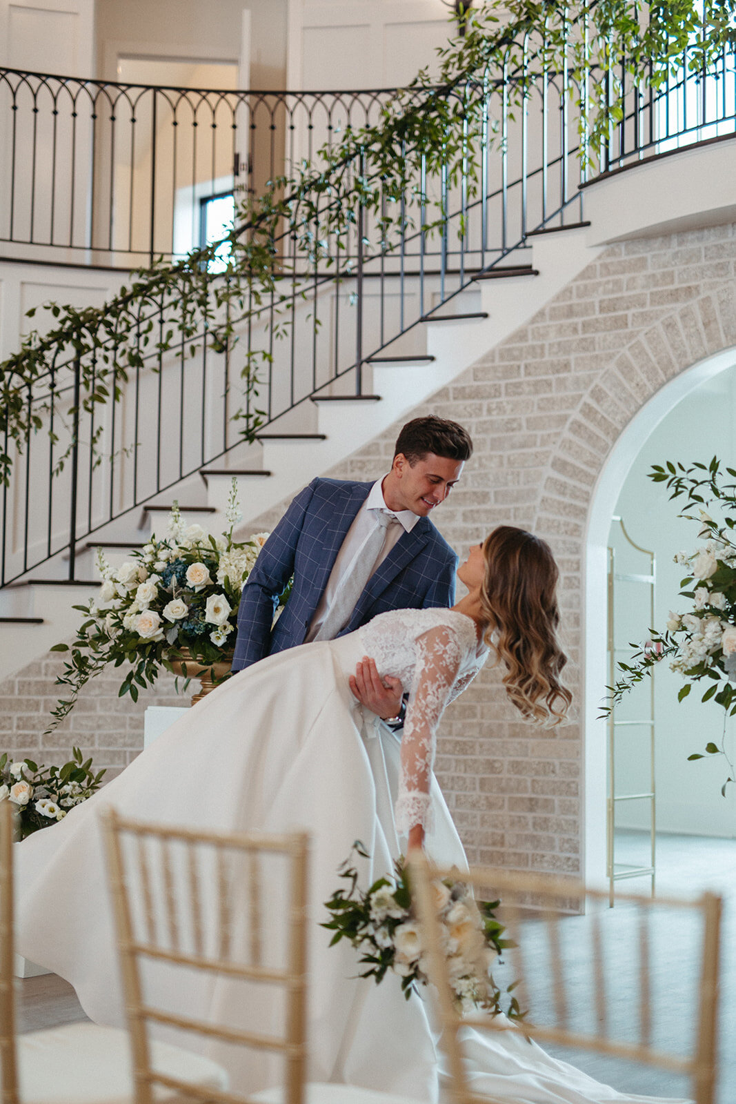 A groom in a blue suit dips his bride wearing a white wedding gown in front of an arched brick entryway with flowers.