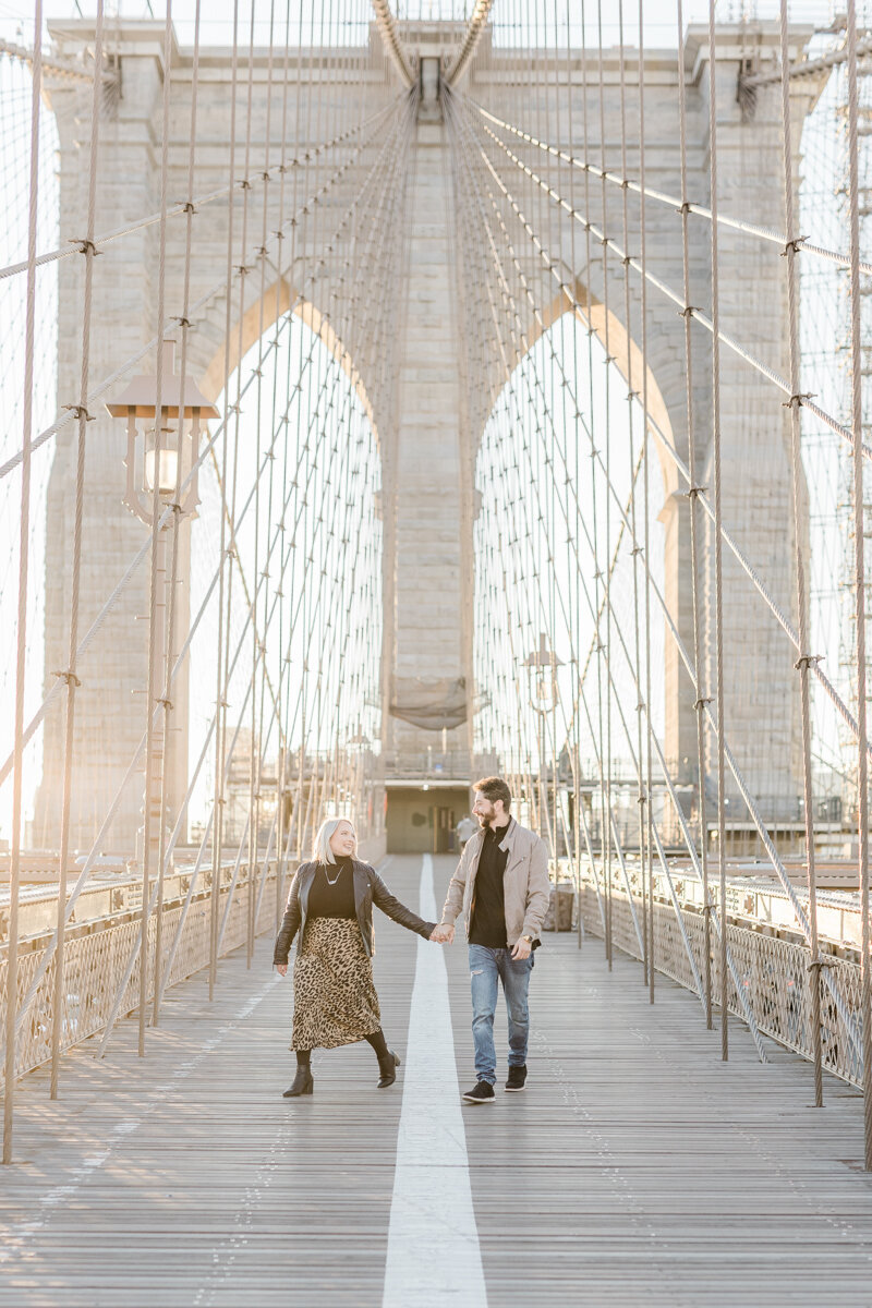 Engaged couple walking hand in hand across the Brooklyn Bridge at sunrise in NYC