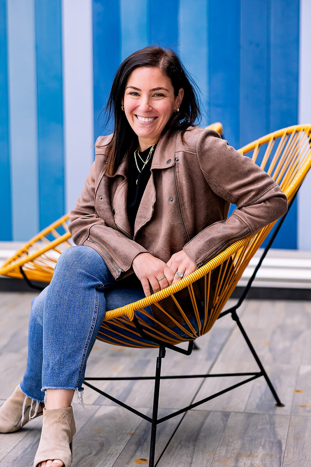 Becky smiling and sitting in a yellow chair while wearing a brown leather jacket