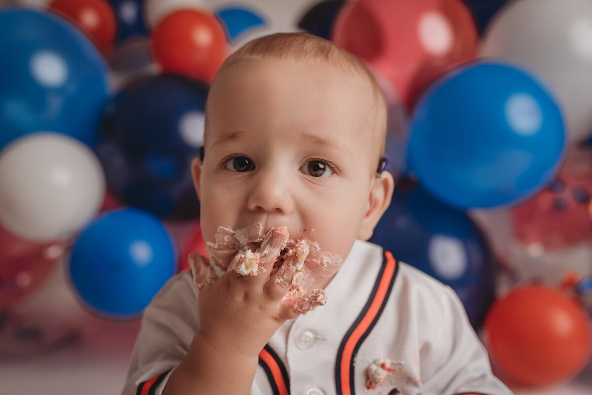 One year old baby boy sitting on floor eating birthday cake with Atlanta Braves baseball jersey on and red, white, blue balloon garland behind him